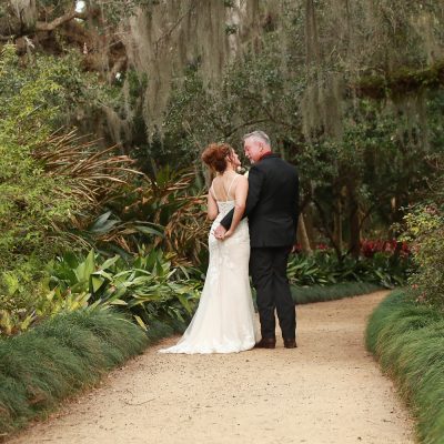 Bride and groom posing on a garden path at Washington Oaks Gardens State Park, surrounded by blooming flowers and tall trees.