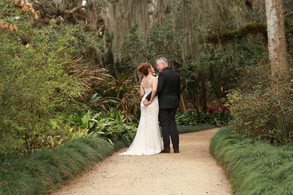Bride and groom posing on a garden path at Washington Oaks Gardens State Park, surrounded by blooming flowers and tall trees.